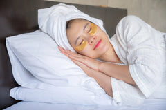 Woman relaxing with gold eye patches and towel wrap, resting peacefully in bed.